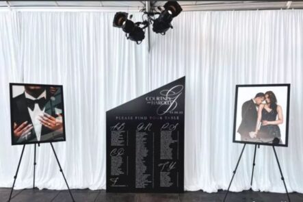 A black seating chart flanked by photos of the bride and groom in front of white drapery