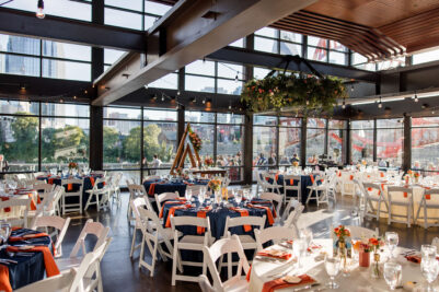 A wedding reception set up in the Cumberland space with have blue and white round tables with white chairs and bright orange napkins with colorful centerpieces in small vases