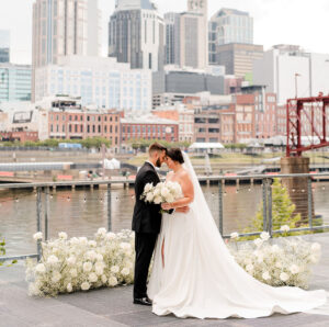 A bride and groom pose with their foreheads touching in front of baby's breath ground arch on the riverfront patio