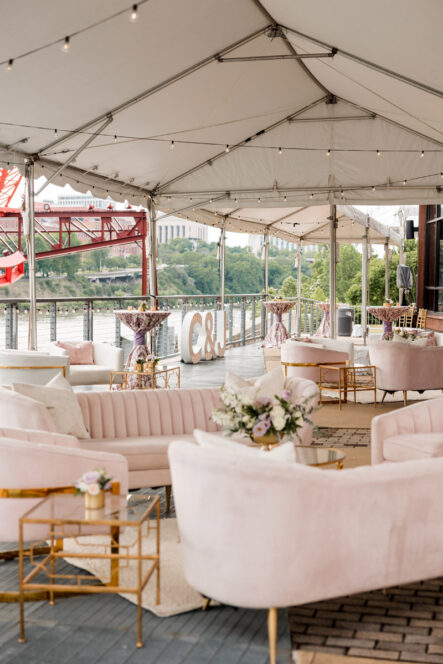 A tented riverfront patio with soft pink lounge furniture for a wedding reception cocktail hour