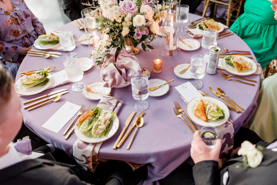 A look down at a reception table with lavender linens and a large floral centerpiece with guests at their seats in front of a plated salad