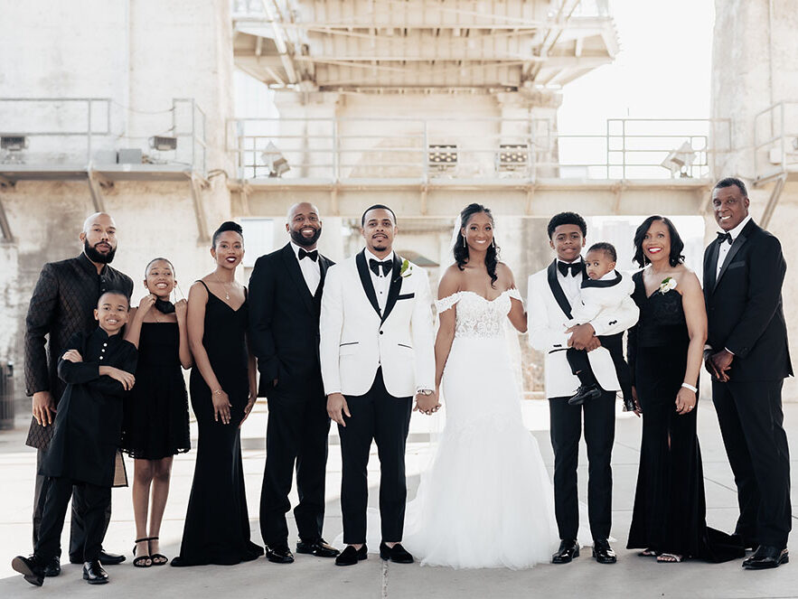 A groom in a white tuxedo holds hands with his bride in an off-the-shoulder gown, posing with their family underneath the pedestrian bridge