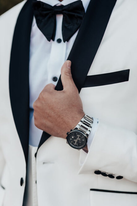 A close-up of a groom's hand holding his white tuxedo's black lapel