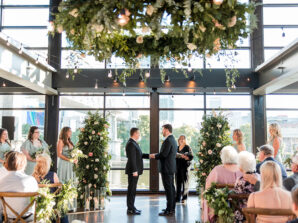 Two grooms hold hands at the alter as guests look on. The ceremony is decorated with a hanging greenery ring with Edison bulb lighting and freestanding greenery arrangements at the altar