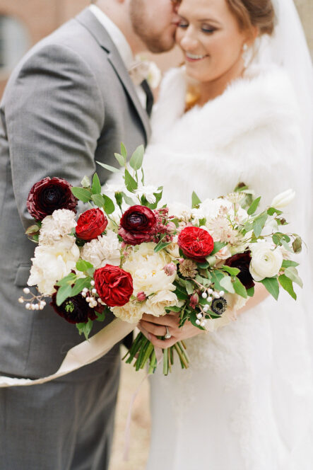 Groom kisses bride's cheek as she smiles in background out of focus with red and white bridal bouquet in foreground for romantic winter wedding at The Bridge Building, downtown Nashville wedding venue