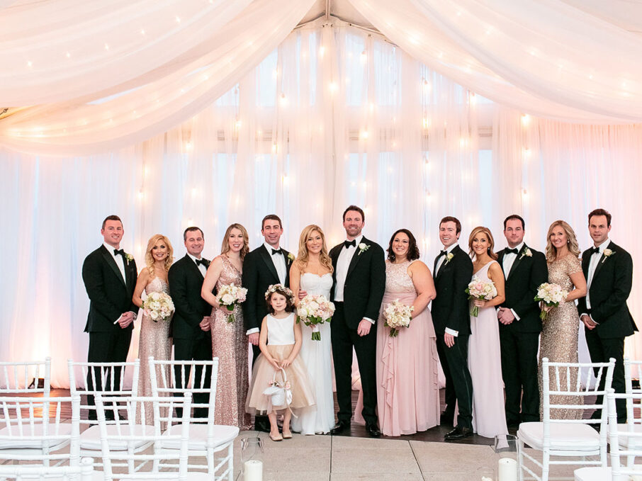 Bride and groom pose with wedding party at ceremony altar before tented wedding ceremony with string lights and white drapery with soft pink lighting for rose gold New Year's Eve wedding at the Bridge Building, a downtown Nashville wedding venue