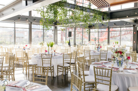Wedding reception setup with greenery hanging from ceiling, white table linens, and light wood chiavari chairs