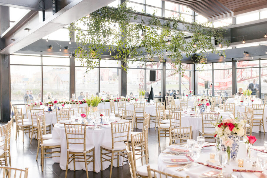 Wedding reception setup with greenery hanging from ceiling, white table linens, and light wood chiavari chairs