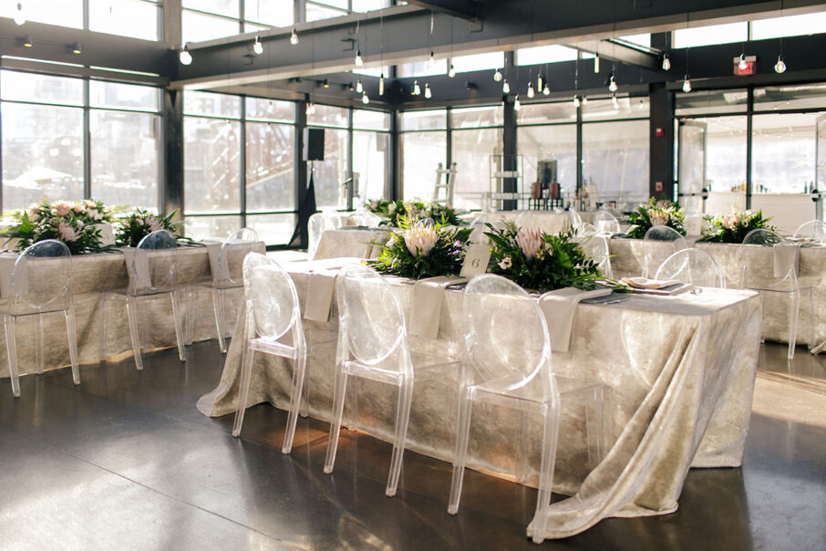 Wedding reception table with ghost chairs, silver textured linens, and large tropical centerpieces with palm leaves and protea flowers for tropical-inspired wedding at the Bridge Building, a downtown Nashville wedding venue