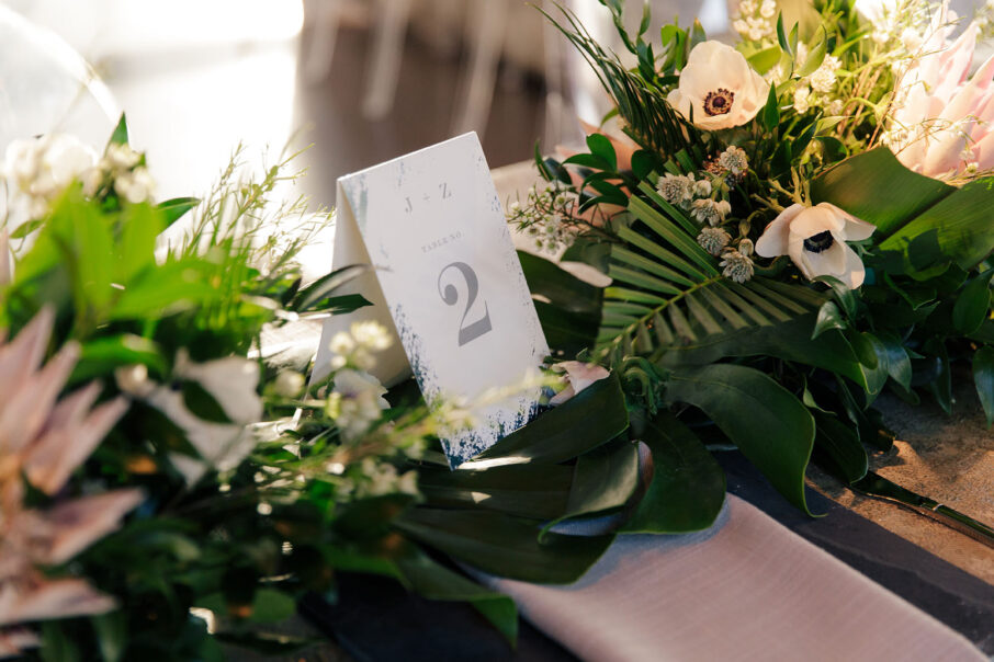A close-up of a blue and silver table number nestled into tropical leaves and flowers at head table for tropical-inspired wedding at the Bridge Building, a downtown Nashville wedding venue