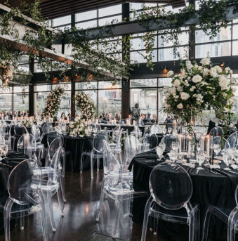 The Cumberland space set up with a grand wedding reception, filled with round tables in black linens, clear acrylic ghost chairs, large white floral centerpieces, a white floral ring hanging at the center, and abundant greenery wrapping the industrial beams of our Nashville wedding venue