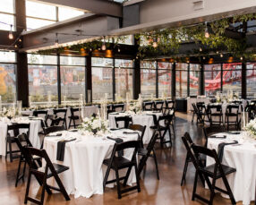 A wedding reception dinner in the Cumberland space with black chairs, white round table linens, black napkins, white centerpieces, and greenery with Edison bulbs hanging from the industrial beams
