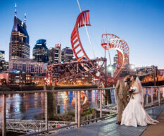 A bride and groom kiss on the riverfront patio overlooking the Cumberland River and the Nashville skyline at dusk.