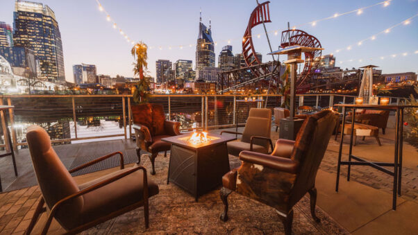 Modern western-inspired lounge furniture surrounding a fire pit underneath string lights on the riverfront patio overlooking downtown Nashville at dusk