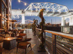 Lounge areas with fire pits set up on the riverfront patio with winter pine decor, overlooking the lit up pedestrian bridge