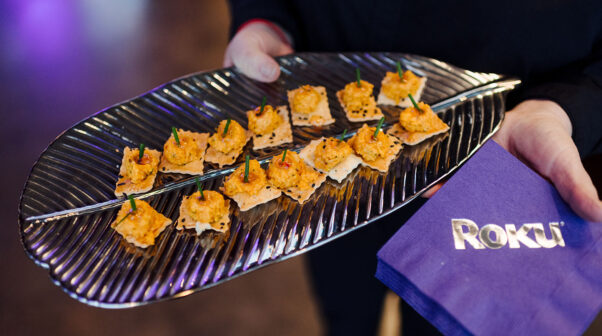 A close-up of a cater waiter holding a plate of pimento cheese bites and purple branded napkins for Roku